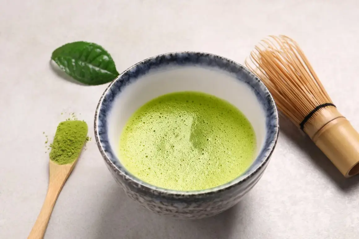 Bol rempli de matcha mousseux, une cuillère en bois avec de la poudre verte et un fouet en bambou. Une feuille de thé verte repose à côté, évoquant le pouvoir antioxydant du matcha.