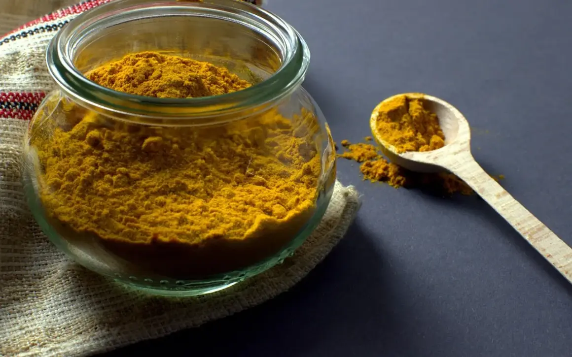 Un pot en verre rempli de poudre de curcuma, avec une cuillère en bois à côté. Ces épices, comme le cumin, apportent couleur et saveur.