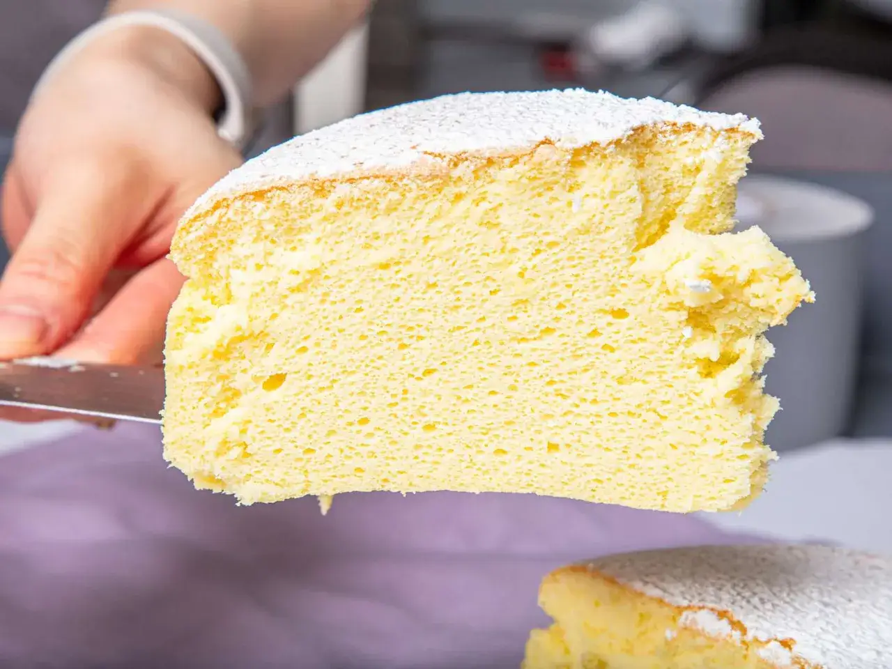 Une main coupe une tranche a&eacute;rienne de cheesecake japonais saupoudr&eacute; de sucre glace. Sa texture l&eacute;g&egrave;re et moelleuse est irr&eacute;sistible.