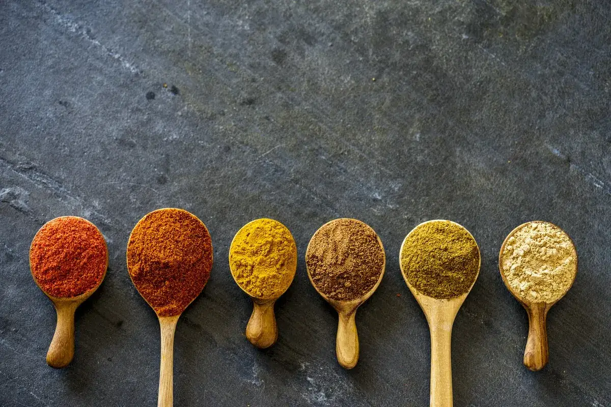 Six cuillères en bois alignées sur une surface sombre, remplies d'épices colorées. Idéal pour une recette de garam masala.