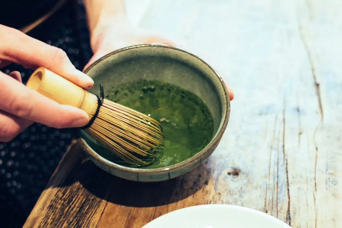 Pr&eacute;paration d'un matcha &agrave; jeun. Une main tient un fouet en bambou pour m&eacute;langer la poudre verte dans un bol.