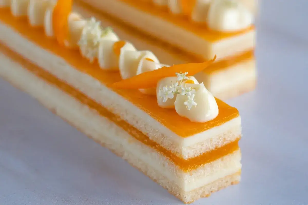 Un délicieux entremet yuzu, aux couches fines et à la garniture crémeuse, orné de zestes d'orange et de petites fleurs blanches.