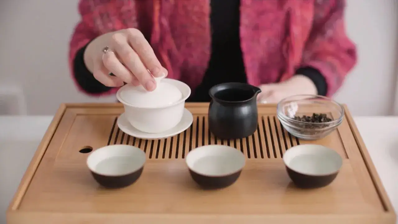 Pr&eacute;paration d'un th&eacute; de Chine traditionnel. Une main soul&egrave;ve le couvercle d'un gaiwan blanc sur un plateau en bambou. Trois tasses sont align&eacute;es.