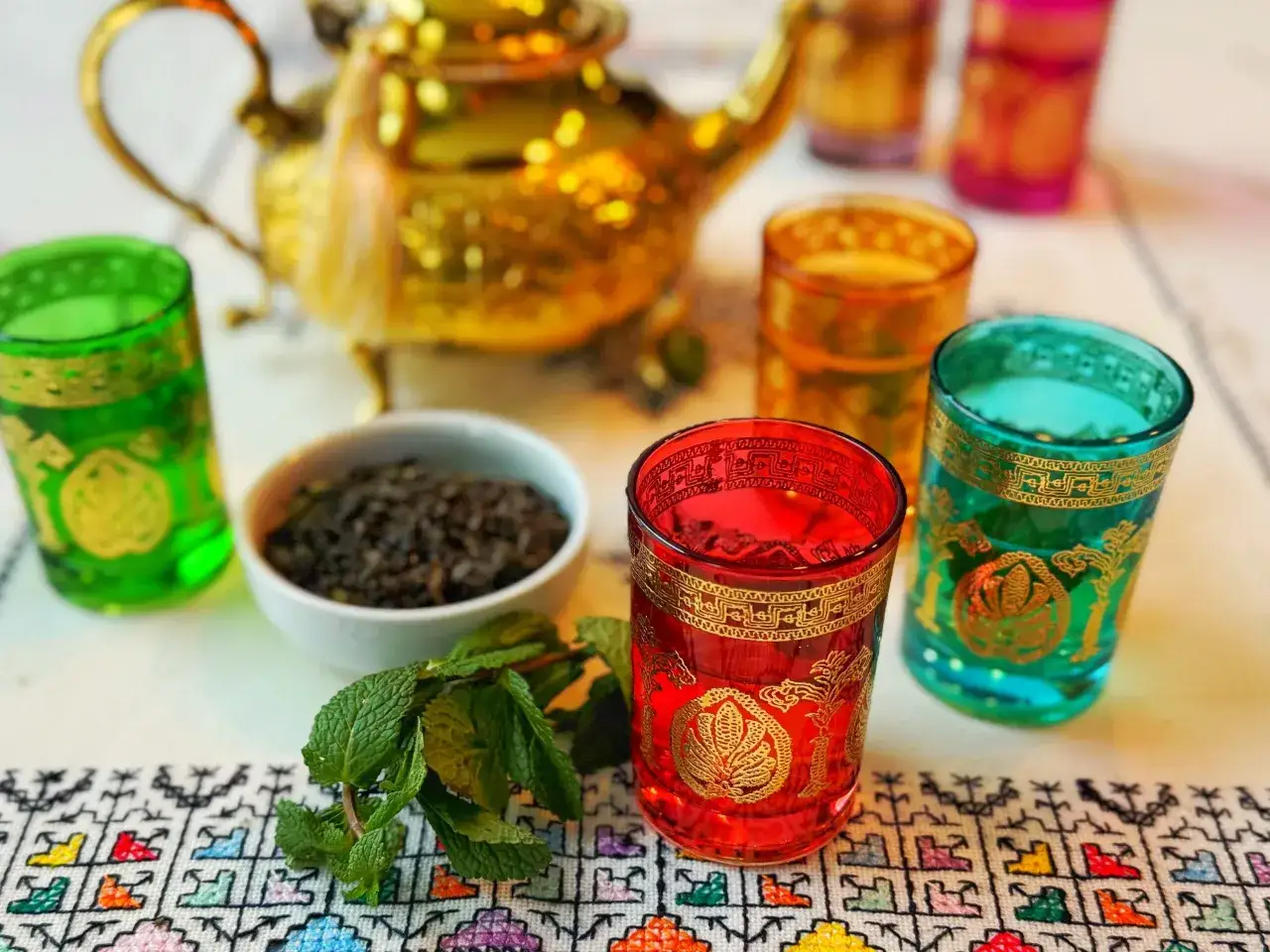 Th&eacute;i&egrave;re dor&eacute;e, verres color&eacute;s et menthe fra&icirc;che sur une nappe brod&eacute;e. Un d&eacute;lice &agrave; la menthe fra&icirc;che.