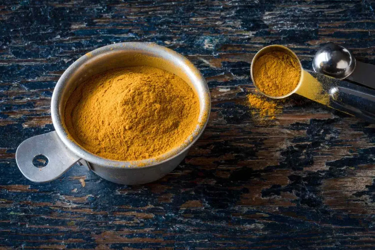 Une cuill&egrave;re doseuse remplie d'&eacute;pice colombo, d'une belle couleur jaune orang&eacute;, repose sur une surface en bois sombre.