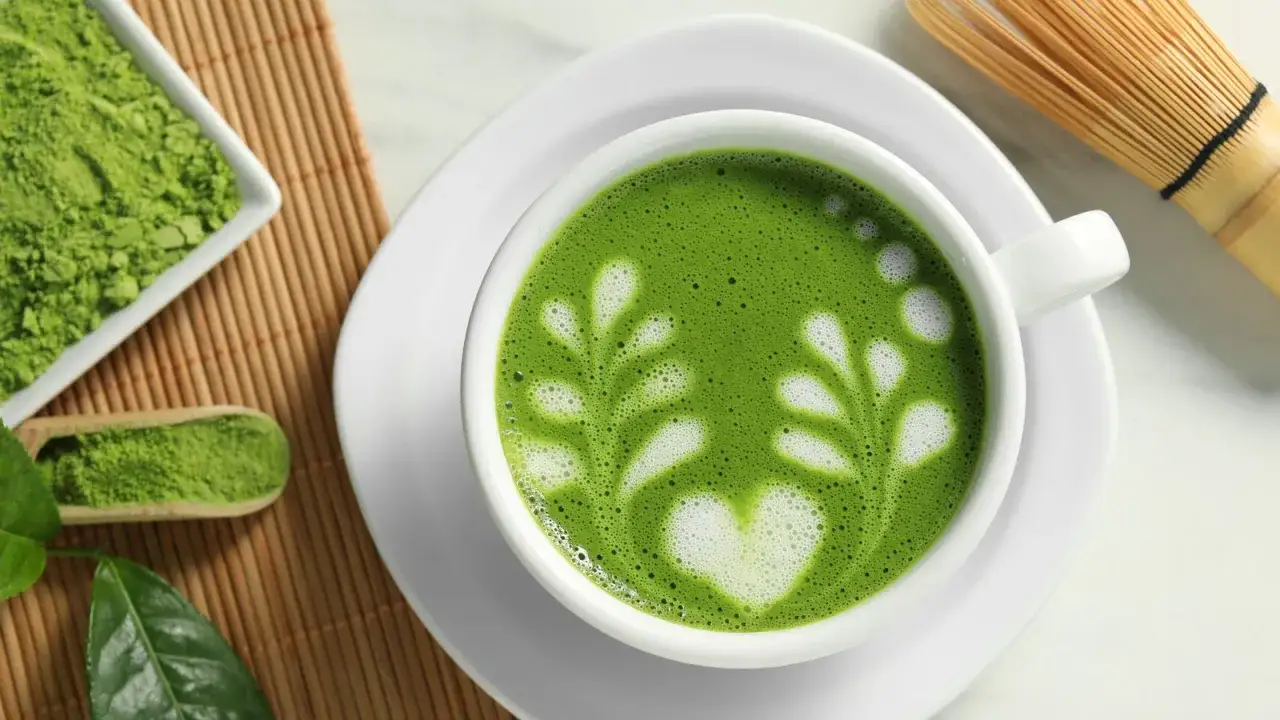 Une tasse de matcha fer, ornée d'un cœur et de feuilles en mousse, à côté de poudre de matcha et d'un fouet traditionnel.