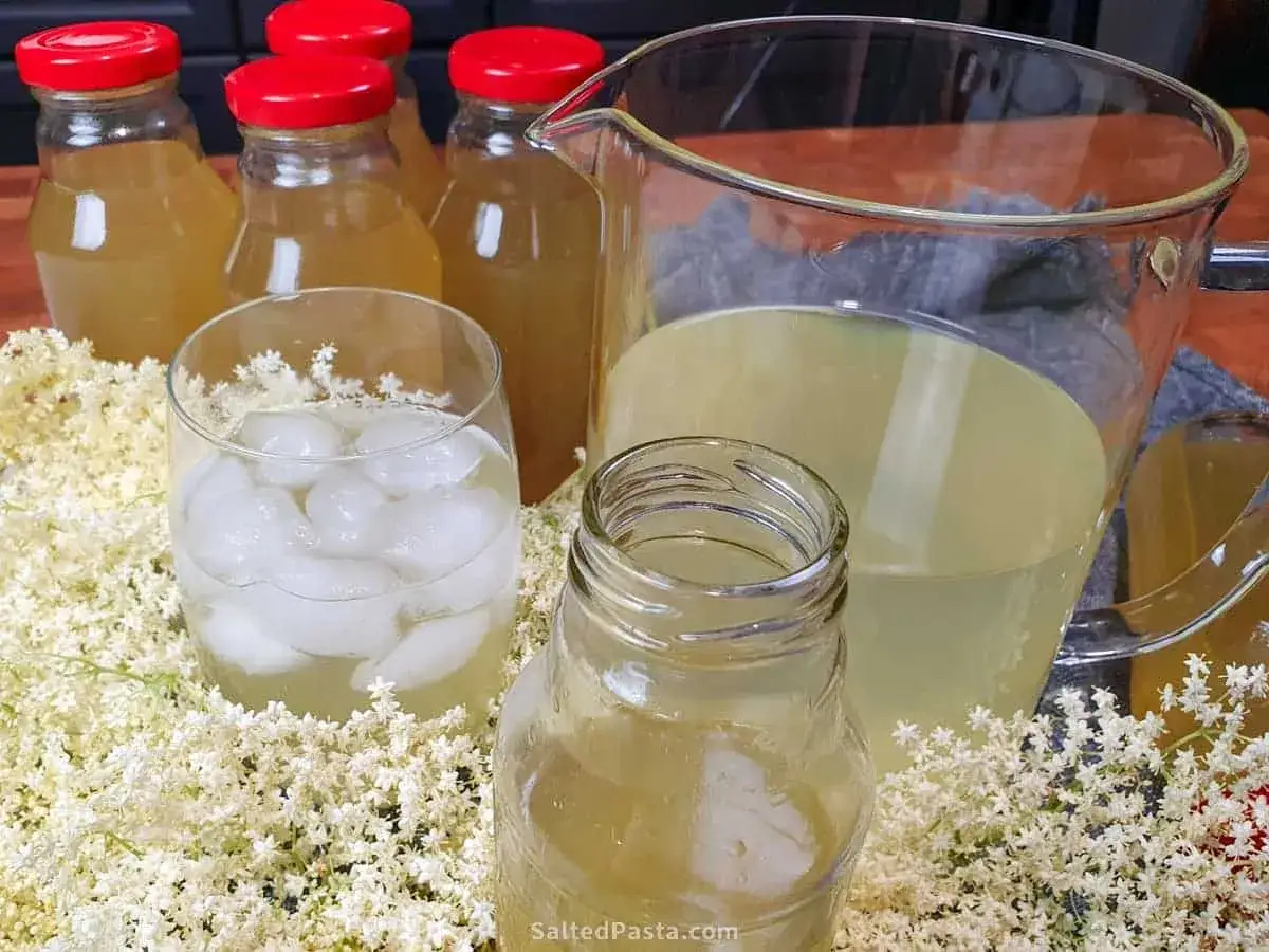 Verre de sirop de sureau glac&eacute;, carafons et pichet, entour&eacute;s de fleurs de sureau. D&eacute;couvrez les bienfaits du sirop de sureau.