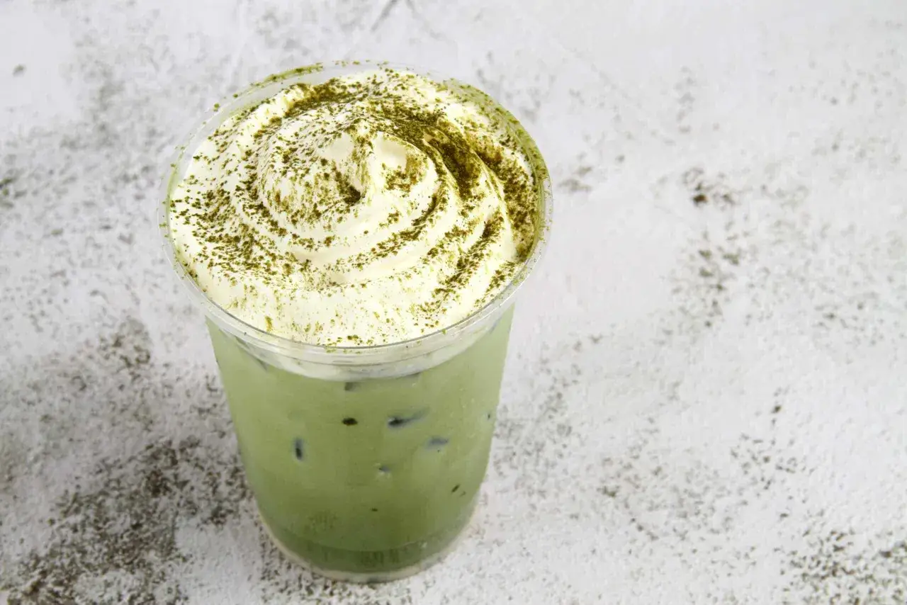 Un gobelet en plastique rempli de matcha glac&eacute;, garni de cr&egrave;me fouett&eacute;e et saupoudr&eacute; de poudre de matcha. Un d&eacute;lice pour ceux qui aiment boire du matcha tous les jours.