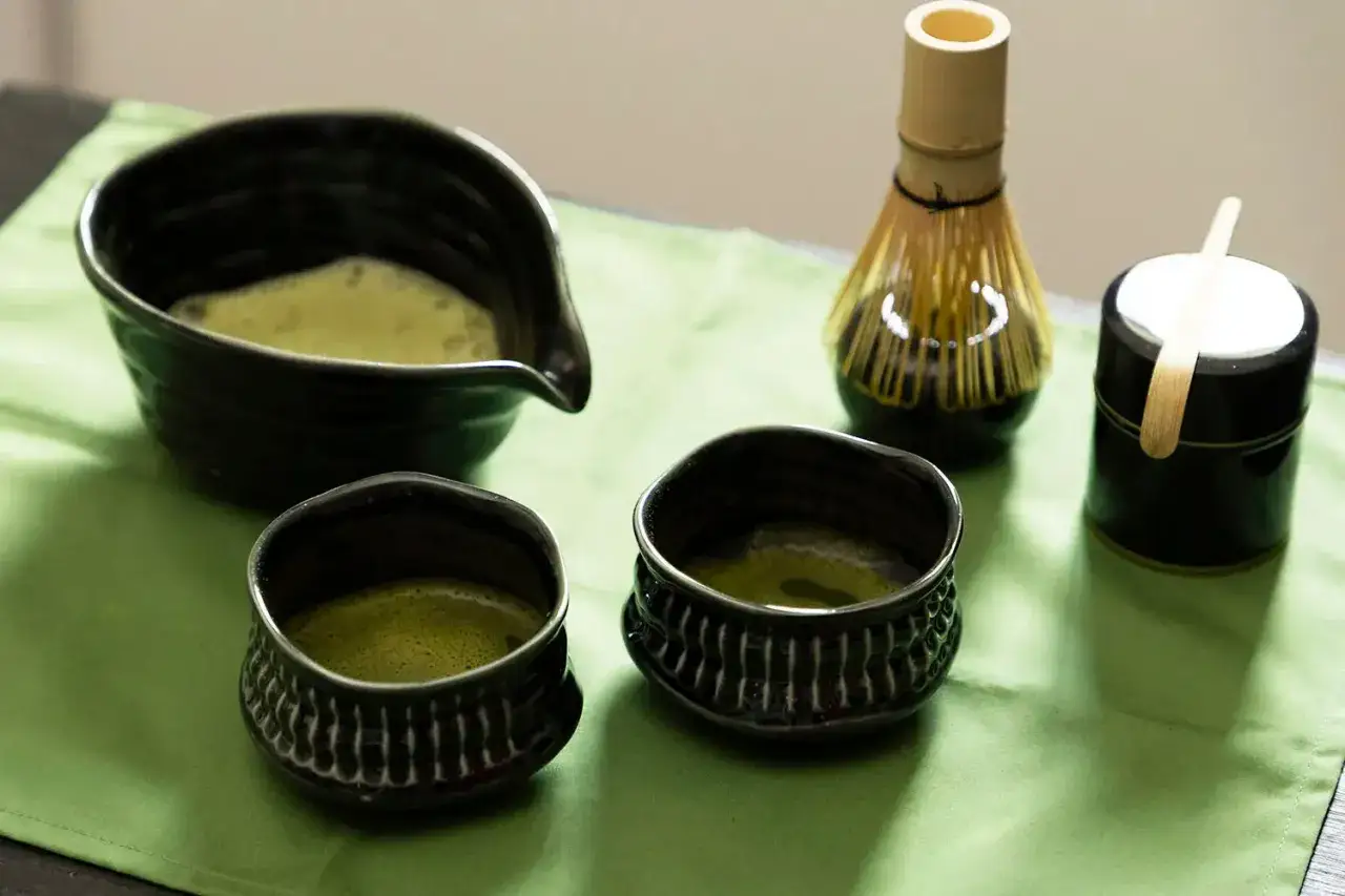 Préparation du matcha : une théière verse le thé vert dans deux tasses. Un fouet et une boîte à matcha sont à proximité, suggérant la quantité de matcha par tasse.
