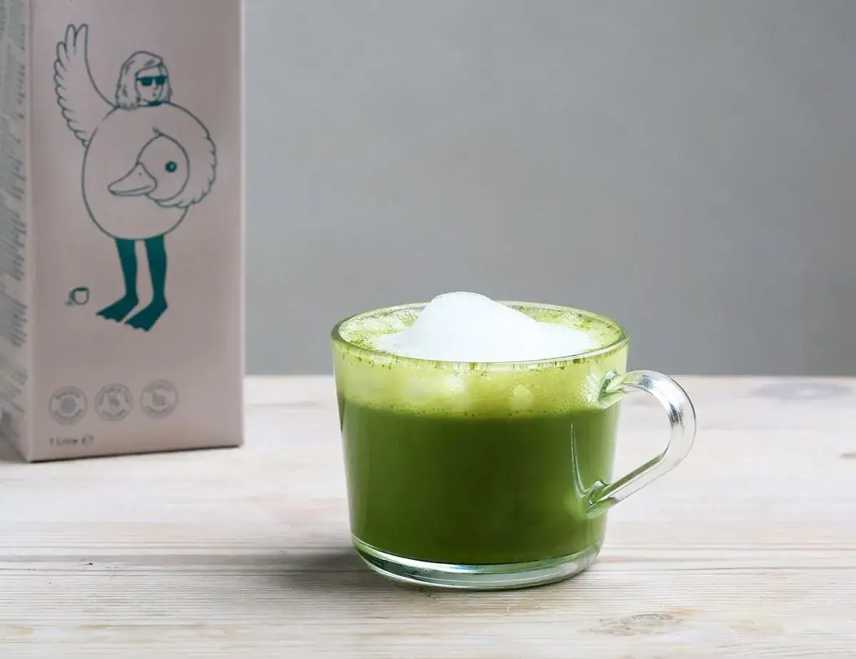 Un matcha lait d'avoine cr&eacute;meux dans une tasse en verre, &agrave; c&ocirc;t&eacute; d'une bo&icirc;te au design original.