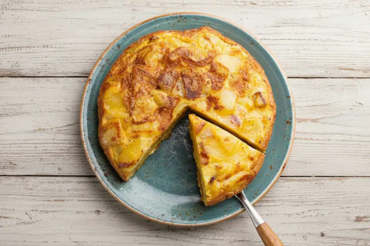 Une d&eacute;licieuse tortilla espagnole, dor&eacute;e et moelleuse, est servie sur une assiette bleue. Une part est d&eacute;j&agrave; coup&eacute;e.