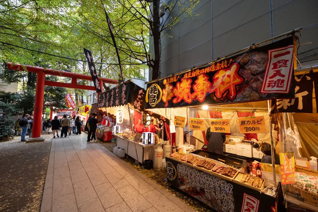 Ambiance de fête avec des stands de street food japonais, des brochettes grillées et des bannières colorées sous un torii rouge.