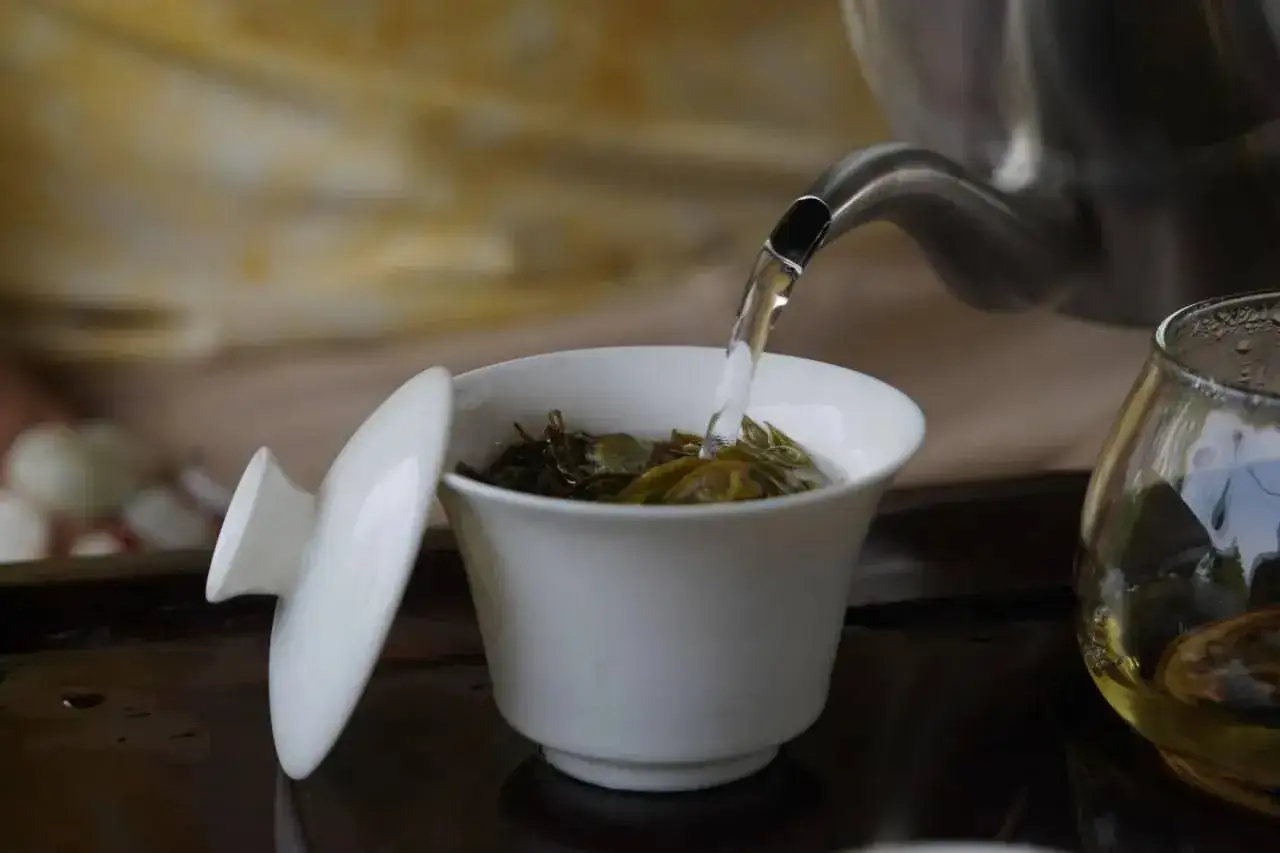 Infusion de thé sencha, révélant ses bienfaits pour la santé. L'eau chaude coule d'une théière dans une tasse blanche remplie de feuilles de thé.