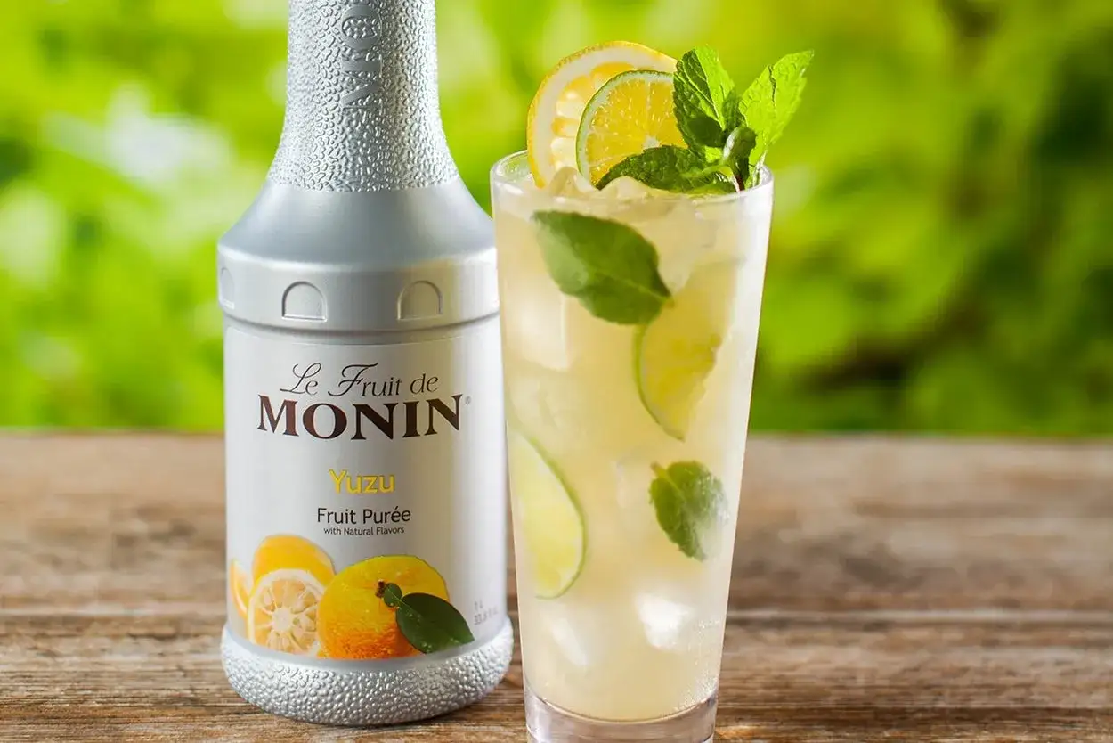 Bouteille de sirop de yuzu Monin et un verre de boisson rafra&icirc;chissante avec des tranches de citron, de lime et de la menthe. Parfait pour une recette de sirop de yuzu maison.