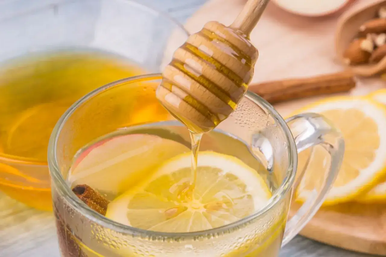 Une cuill&egrave;re en bois verse du miel dans une tasse de boisson chaude aux citrons, pommes et cannelle, &eacute;voquant les bienfaits de la cannelle du miel et du citron.