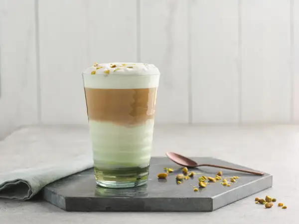 Un latte pistache aux couches distinctes, couronné de mousse et de pistaches concassées. Une cuillère en cuivre repose à côté.