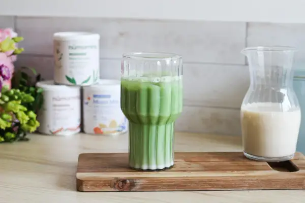 Un verre de matcha chaud, mousseux et crémeux, prêt à être dégusté. À côté, une carafe de lait végétal.