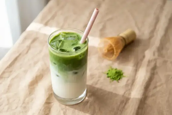 Un verre de pâte minute claire au matcha, glacé et rafraîchissant, avec une paille rose. Un fouet à matcha et une pincée de poudre verte à côté.