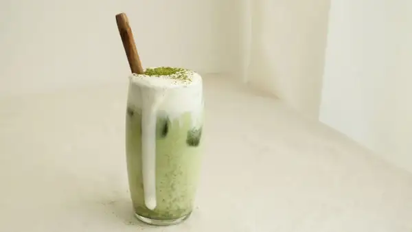 Un verre de matcha lait d'amande crémeux, garni de mousse et d'une pincée de poudre verte, avec une cuillère en bois.