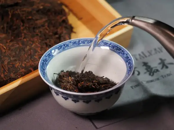 Préparation d'un thé de Chine. De l'eau chaude est versée dans un bol rempli de feuilles de thé sombres, à côté d'un bloc de thé compressé.
