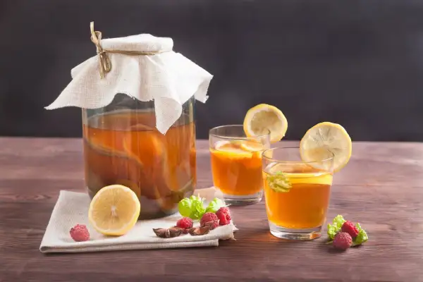 Un grand bocal de boisson kombucha, deux verres garnis de citron et de menthe, avec des framboises et une tranche de citron sur une serviette.