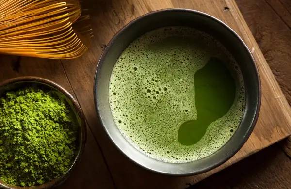 Une composition de matcha : poudre verte vibrante dans un bol, une tasse de thé mousseux et un fouet en bambou sur fond de bois.