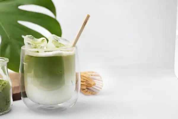 Un verre de matcha à jeun, glacé et crémeux, avec une paille en bois. À côté, un fouet à matcha et une feuille de Monstera.
