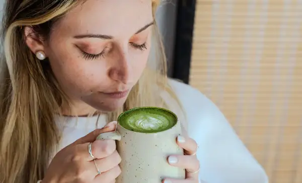 Femme aux cheveux blonds savourant un matcha latte crémeux, le regard clos. La boisson verte présente un joli dessin de mousse.