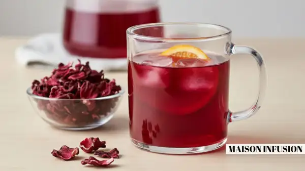 Verre de boisson rouge vif avec glaçons et tranche d'orange, à côté d'un bol de fleurs d'hibiscus séchées. L'eau d'hibiscus, rafraîchissante et colorée.