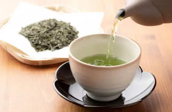 Comment faire du thé vert : une théière verse un liquide vert vif dans une tasse, à côté d'une pile de feuilles de thé séchées.